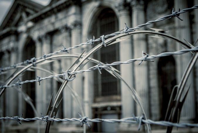 Spirals of razor wire and barbed wire limit access to one of Detroit's abandoned buildings—Michigan Central Station. The focus in the image is on the razor wire and the image has a dark and desaturated tone. The large columns and detailed architecture of the building are visible behind the fence.